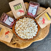 A wood bowl with6 oz bags of Omnivore SALT, LIMONE, PORCINI Salt blends and bags of Ranho Gordo Crimson popping corn with a bowl of pop corn in the center.