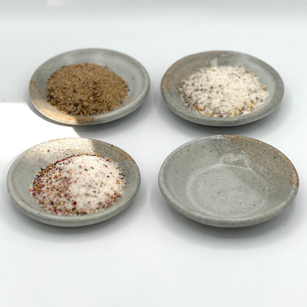 This photo has 4 hand crafted small, white and grey ceramic dishes. 3 of the dishes hold Omnivore SALT, LIMONE and Porcini sea salt blends.